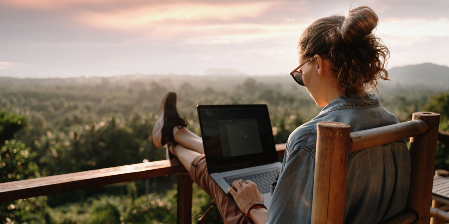 A woman in front of her laptop sitting on a deck with a landscape view.